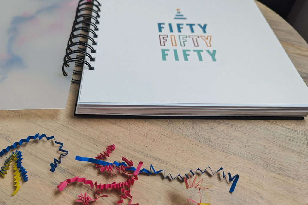 Notebook with 'Fifty Fifty Fifty' text on a wooden surface with colorful birthday confetti.