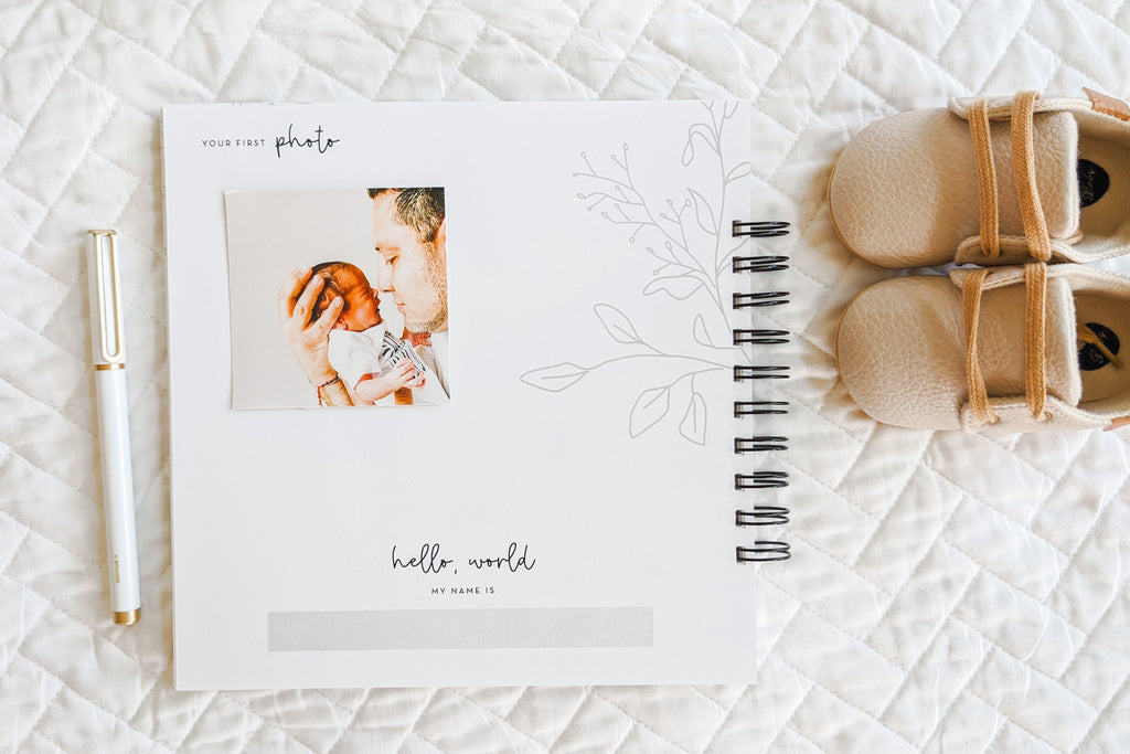 Your first photo page with photo of baby and dad inside pregnancy journal book to track pregnancy week by week with pictures of baby and belly and journaling space sitting on white quilt with baby shoes