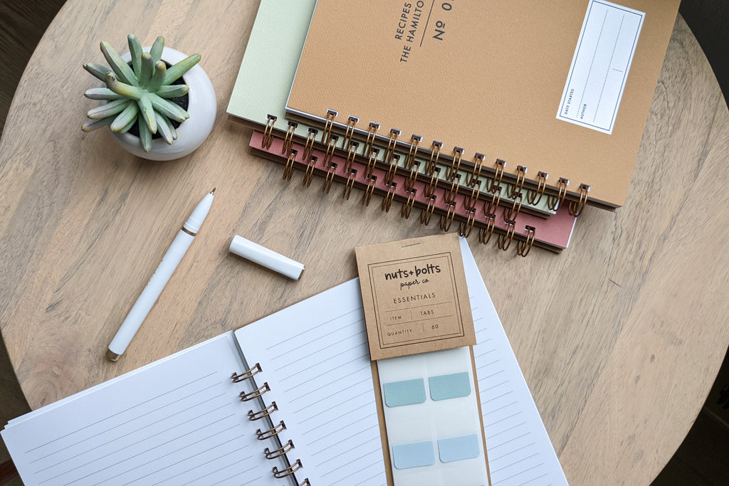 The Vintage Minimalist notebook in several colors including rose, champage, denim and pistachio with a customizable cover that reads whatever you'd like and wire spiral bound sitting on a wooden table with a white pen and an open notebook with lined pages