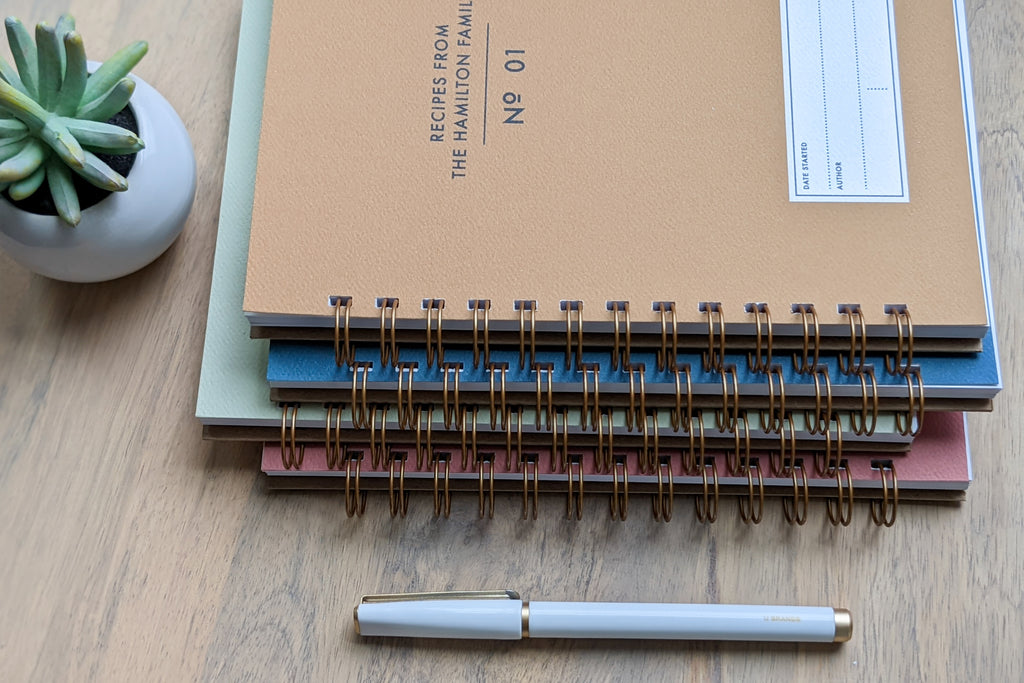 The Vintage Minimalist notebook in several colors including rose, champage, denim and pistachio with a customizable cover that reads whatever you'd like and wire spiral bound sitting on a wooden table with a white pen