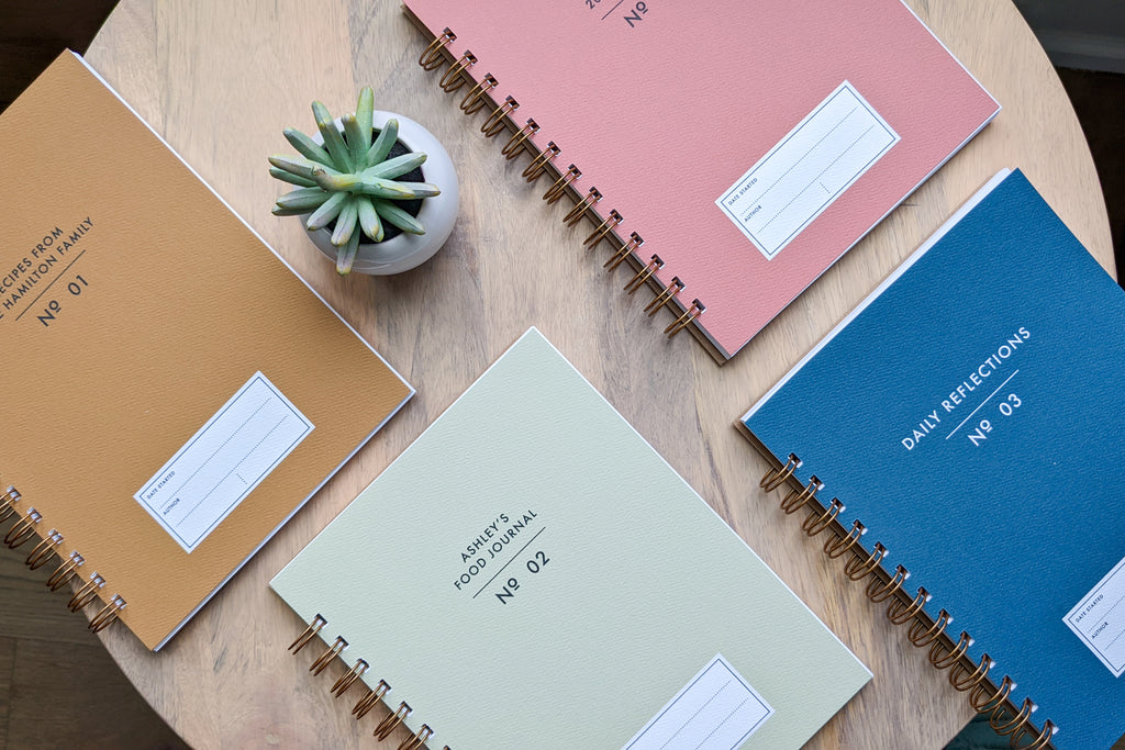 The Vintage Minimalist notebook in several colors including rose, champage, denim and pistachio with a customizable cover that reads whatever you'd like and wire spiral bound sitting on a wooden table with a small plant