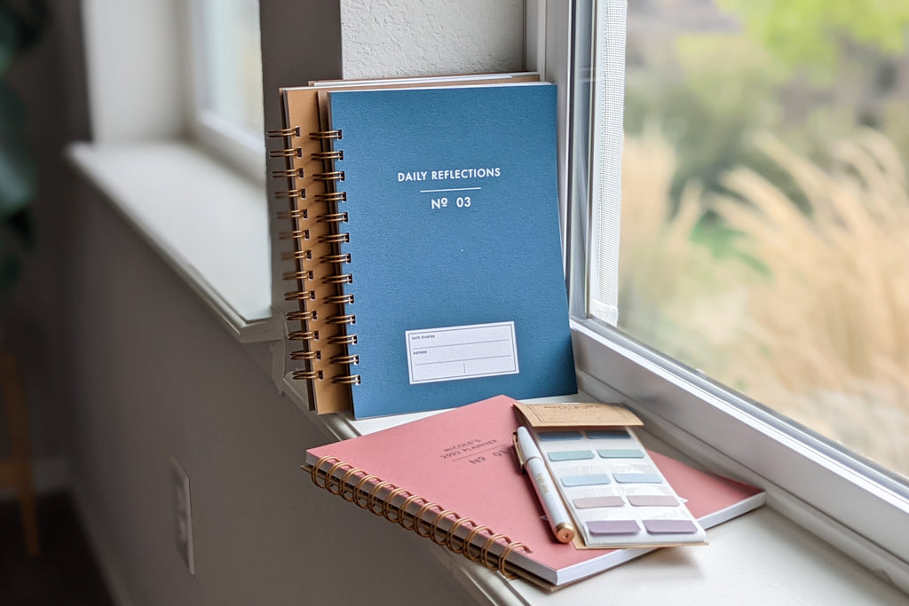 The Vintage Minimalist notebook in several colors including rose, champage, denim and pistachio with a customizable cover that reads whatever you'd like and wire spiral bound sitting on a white windowsill with a white pen and page sticky tabs