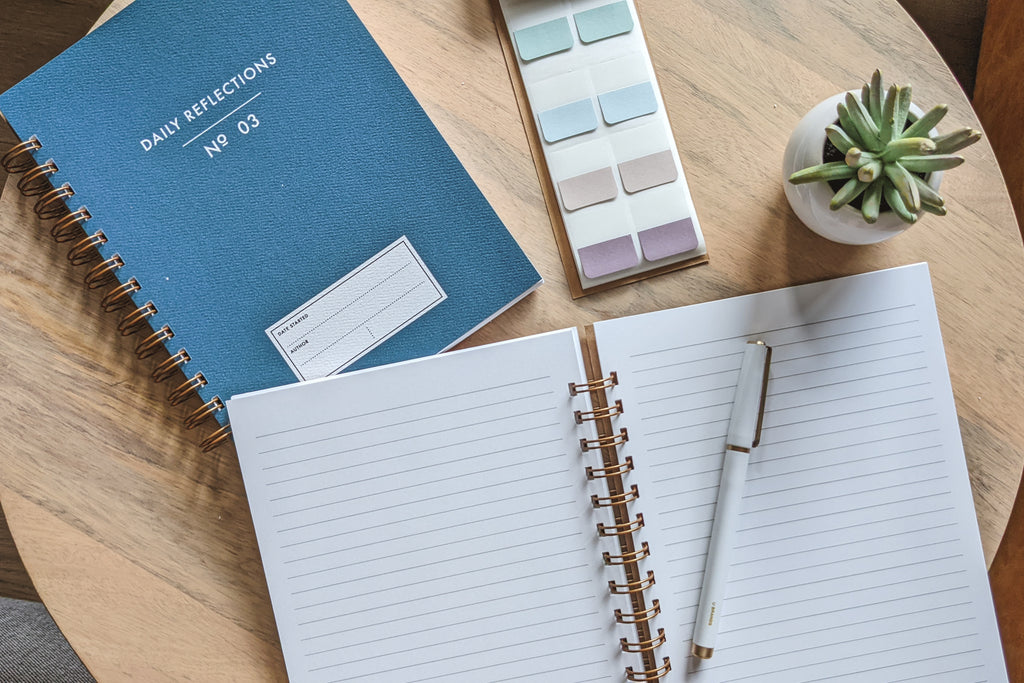 The Vintage Minimalist notebook in several colors including rose, champage, denim and pistachio with a customizable cover that reads whatever you'd like and wire spiral bound sitting on a wooden desk with a white pen, small plant and page tabs