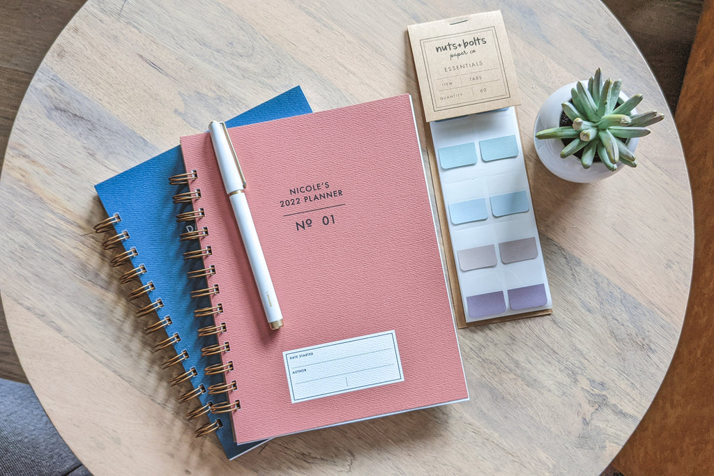 The Vintage Minimalist notebook in rose and denim with a customizable cover that reads whatever you'd like and wire spiral bound sitting on a wooden table with a white pen, small plant and page sticky tabs