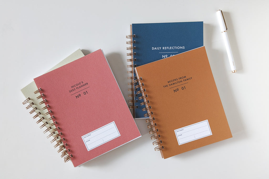 The Vintage Minimalist notebook in several colors including rose, champage, denim and pistachio with a customizable cover that reads whatever you'd like and wire spiral bound sitting on a white desk with a white pen