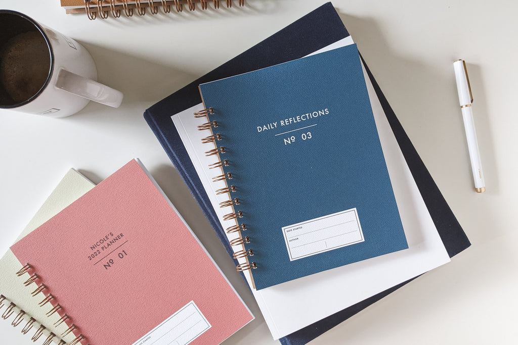 The Vintage Minimalist notebook in several colors including rose, champage, denim and pistachio with a customizable cover that reads whatever you'd like and wire spiral bound sitting on a white desk with a white pen